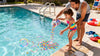 Man and child by a pool, with colorful water balloons being thrown into the water.
